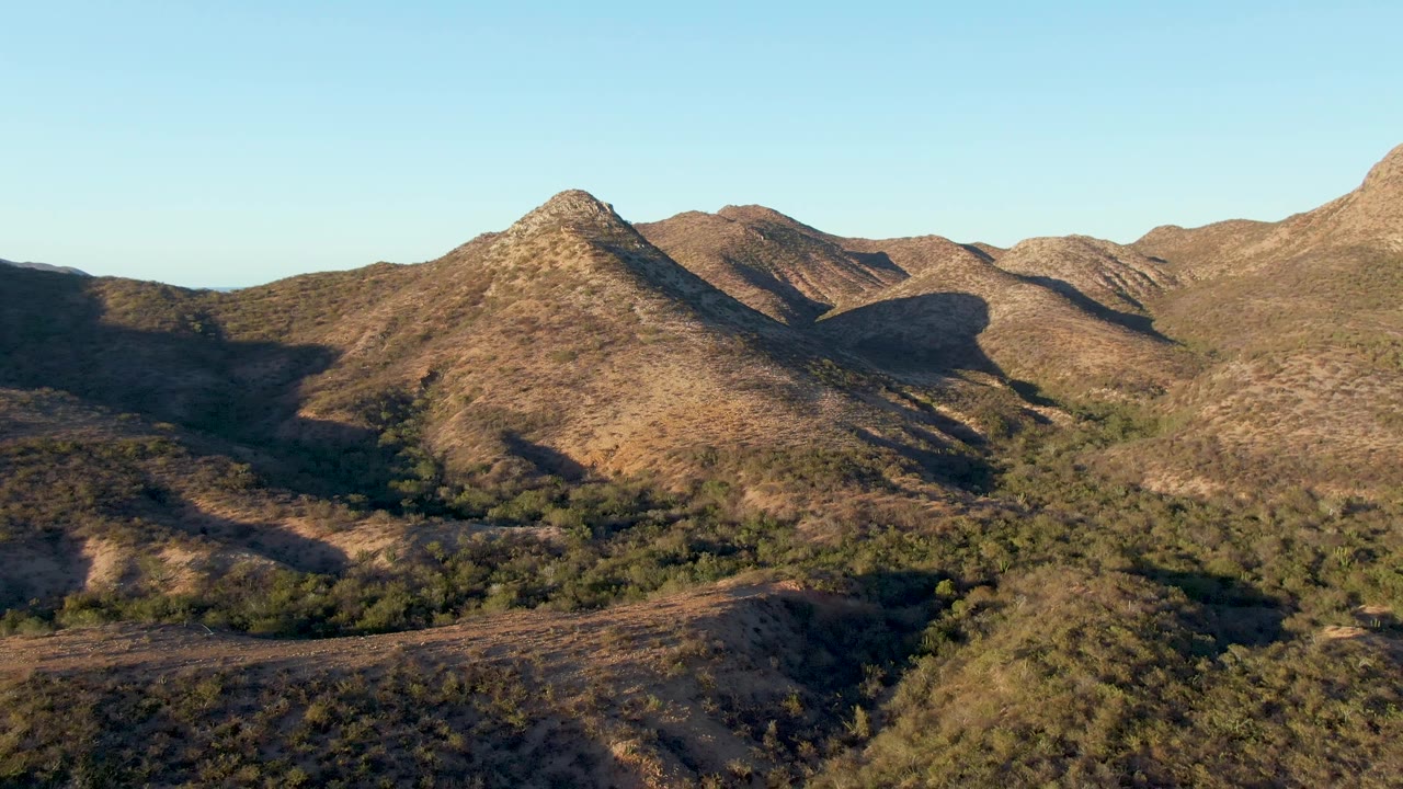 Aerial frame over Cabo Pulmo hills with the central peak shifted slightly left, a broad shadow across the lower terrain, scrubby green patches, distant mountains, and no visible people.