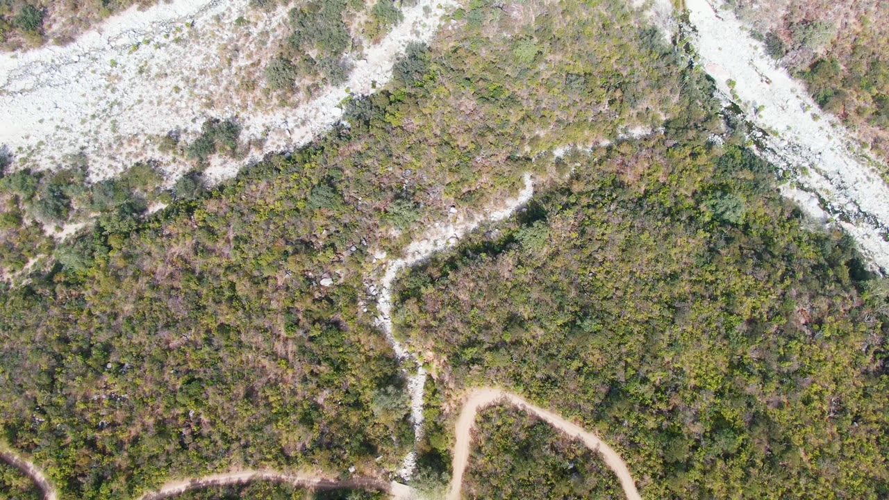 One second into the San Dionisio clip, the overhead view keeps the narrow dirt trail near the lower frame while pale rocky terrain runs across the upper edge, with no visible people.