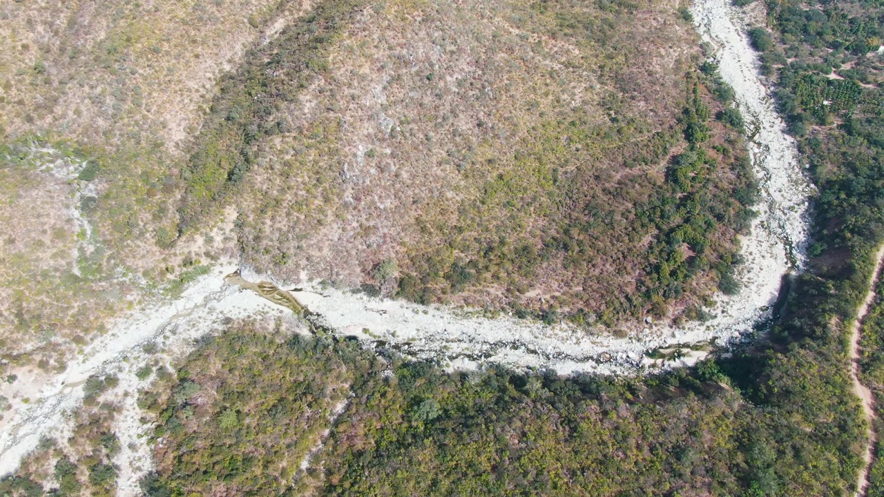 Overhead frame in San Dionisio, Baja California Sur showing the winding pale route crossing mixed dry grass, rocky earth, and green brush, with no visible people.