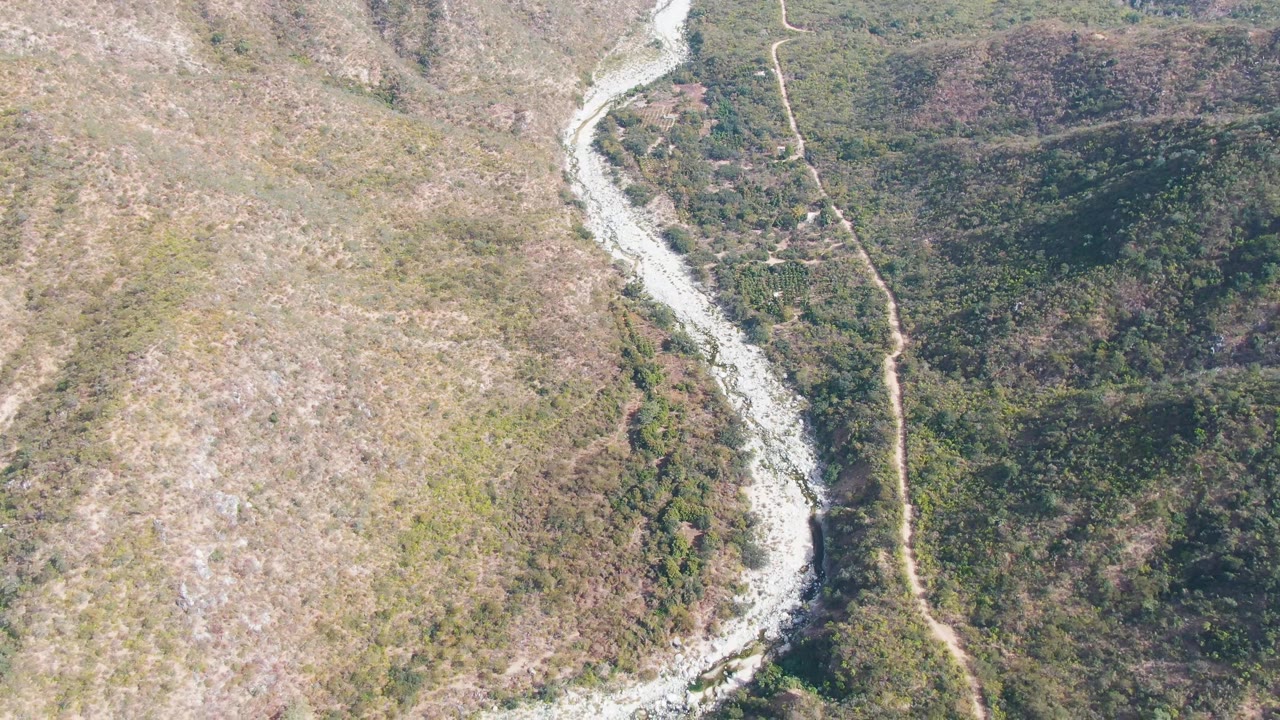 A later aerial frame over San Dionisio shows the river running along the right side of a dry hillside, with denser dark green vegetation across the valley and no visible people.