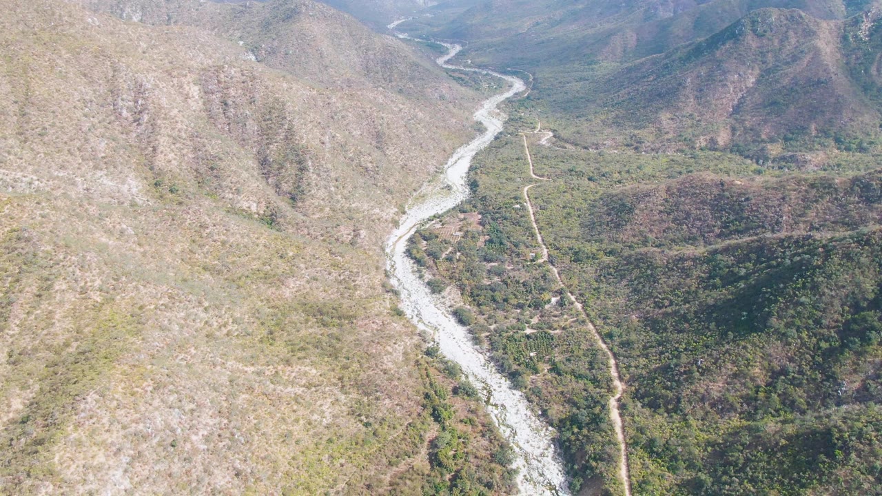 High aerial view over San Dionisio, Baja California Sur, with a pale river curving through a green and brown mountain valley beside a narrow dirt road, no visible people.