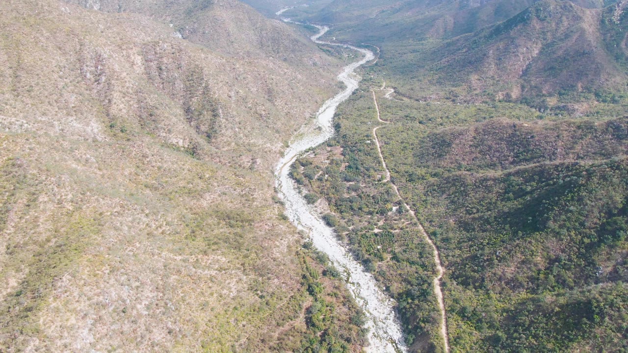 Aerial frame in San Dionisio showing the riverbed bending down the valley while a dirt road tracks alongside brushy green and brown terrain, no visible people.