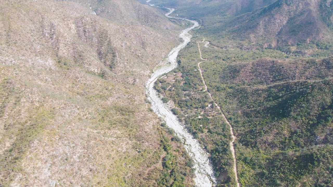 San Dionisio aerial frame with a pale river channel descending through the center of the valley, a dirt road beside it, and mixed green-brown vegetation on the hillsides, no visible people.