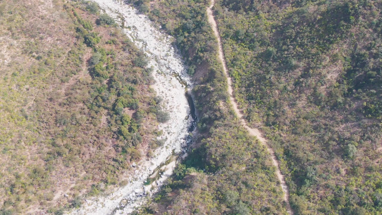 Mid-clip overhead frame from San Dionisio shows the rocky stream bending near dense brush, dry tan ground, and the parallel dirt trail, with no visible people.