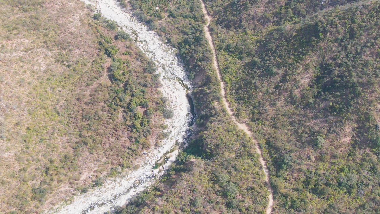 Opening aerial frame in San Dionisio, Baja California Sur, showing a pale rocky riverbed bending through scrub-covered hills with a dirt road beside it and no visible people.