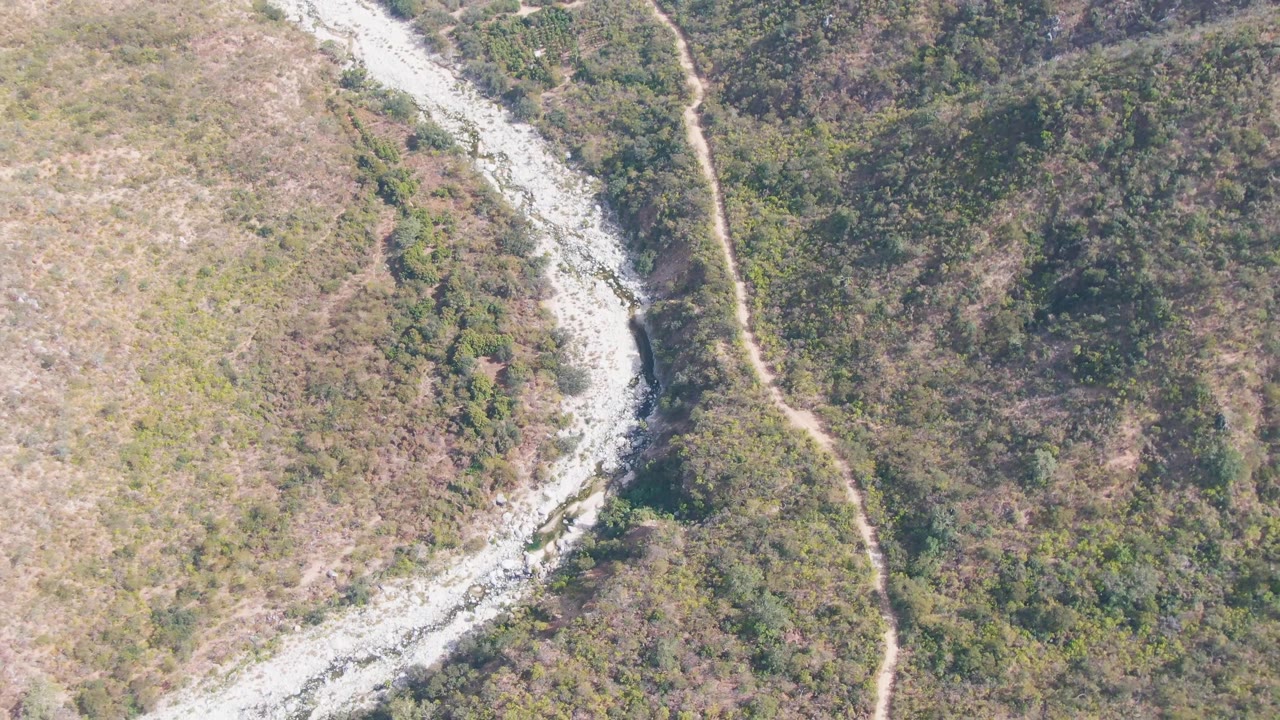 Aerial view in San Dionisio, Baja California Sur, with the river channel shifted slightly through frame and the narrow dirt road tracing the hillside, with no visible people.