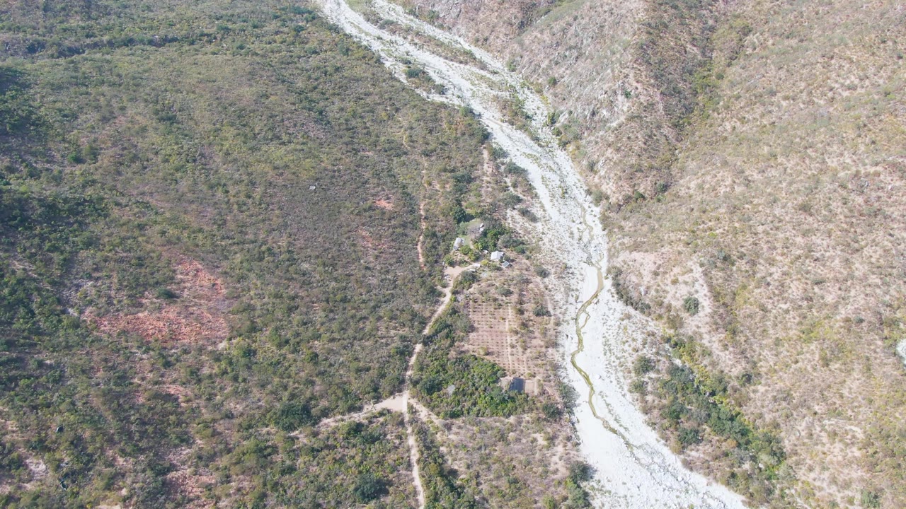 Opening aerial frame over San Dionisio, Baja California Sur, showing a pale winding riverbed cutting between scrub-covered slopes with no visible people.