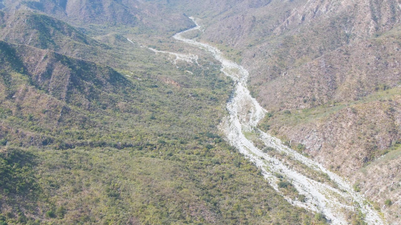 Aerial frame from San Dionisio, Baja California Sur showing a pale riverbed entering from the upper valley and curving through brushy mountain terrain, with no visible people.