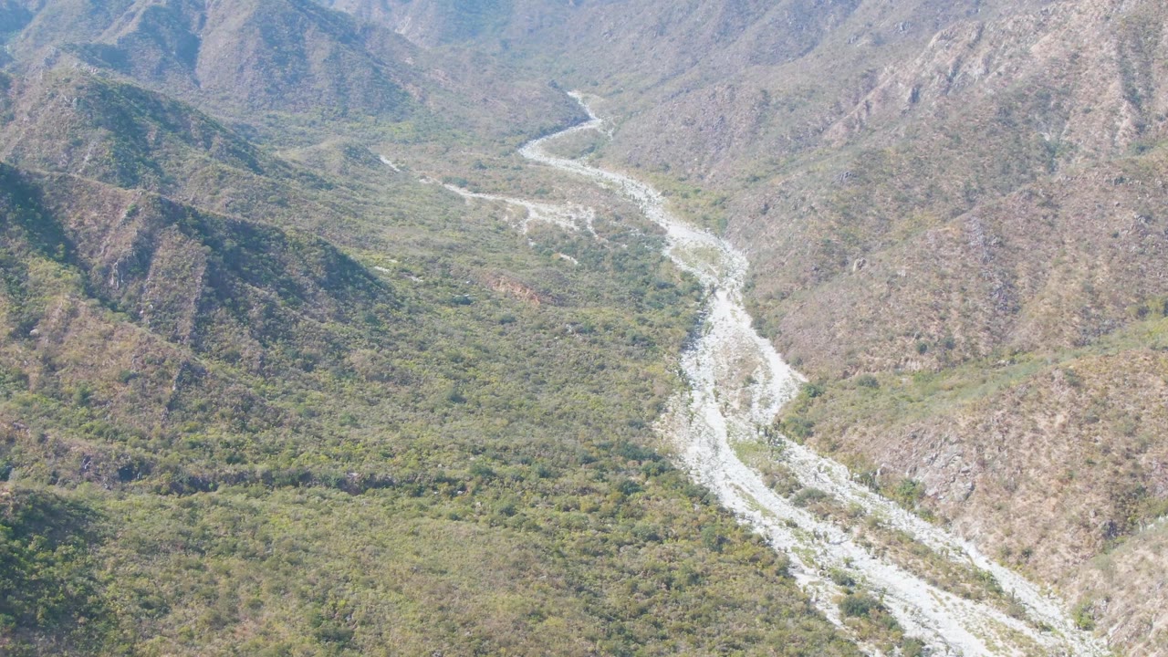Aerial frame from San Dionisio, Baja California Sur showing the pale riverbed splitting across stones beneath steep brown hills and scattered green brush, with no visible people.