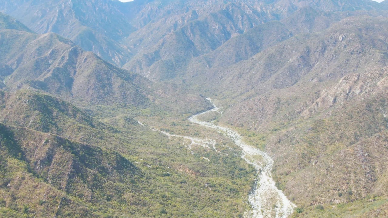 Wide San Dionisio aerial frame showing a bright riverbed snaking away from the foreground into layered Baja California Sur mountains, with no visible people.