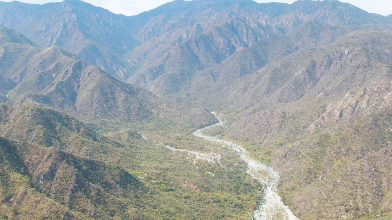 Mid-clip aerial view from San Dionisio showing the winding river lower in frame, green vegetation across the valley floor, and large rocky mountains beyond, with no visible people.