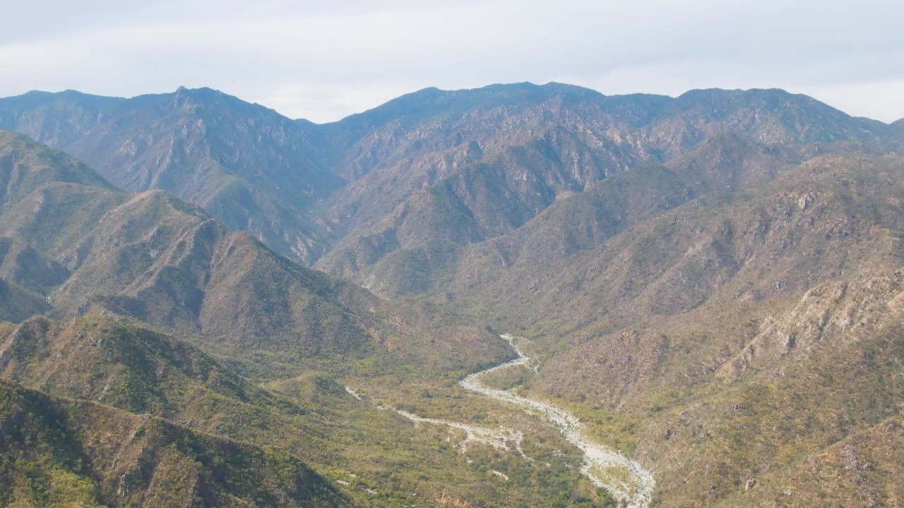 Aerial view in San Dionisio, Baja California Sur, with the riverbed more centered as it threads between rounded green and brown mountain slopes, no visible people.