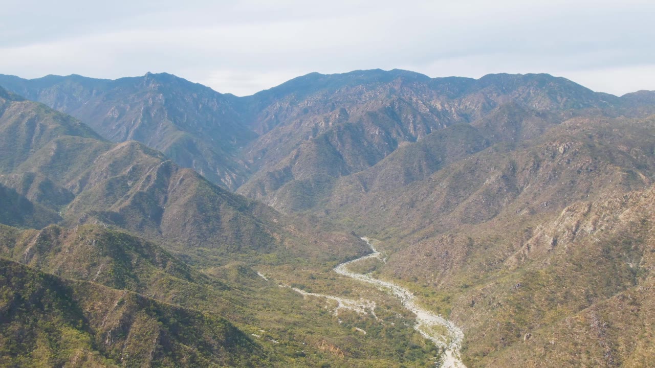 Broad San Dionisio aerial frame showing a pale river course crossing the lower valley beneath rugged ridges and mixed green-brown terrain, no visible people.