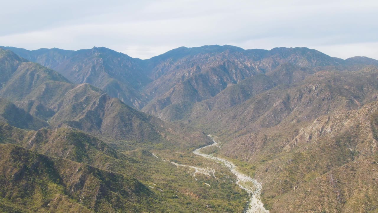 San Dionisio aerial frame with the riverbed entering from the lower left and bending into a dry mountain valley, with layered ridgelines and no visible people.
