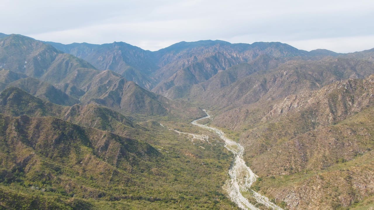 Wide aerial frame over San Dionisio, Baja California Sur, showing a pale winding river through green and brown mountain terrain with no visible people.