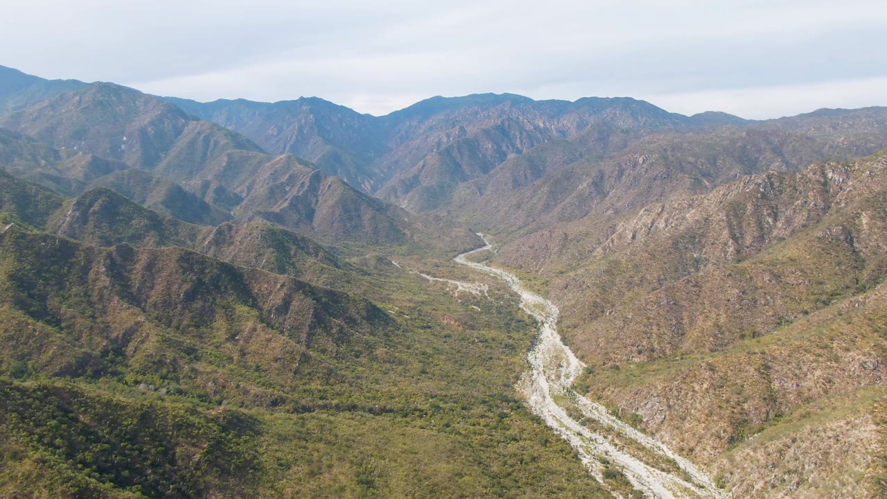High aerial view in San Dionisio, Baja California Sur, with a pale river channel crossing the valley floor below rugged brown-green ridges and no visible people.