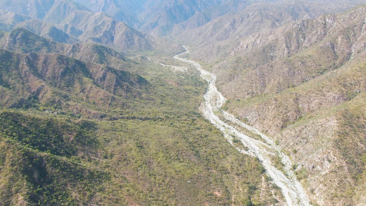 Aerial view over San Dionisio, Baja California Sur, showing a pale winding riverbed between green-brown rocky hills with no visible people.