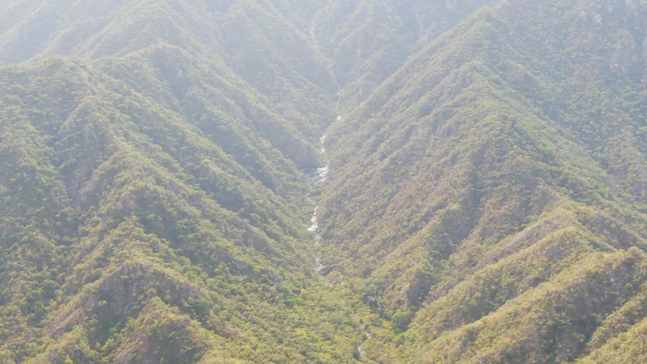 Aerial frame over San Dionisio, Baja California Sur, showing steep green ridges, a narrow central valley, hazy distant peaks, and no visible people.