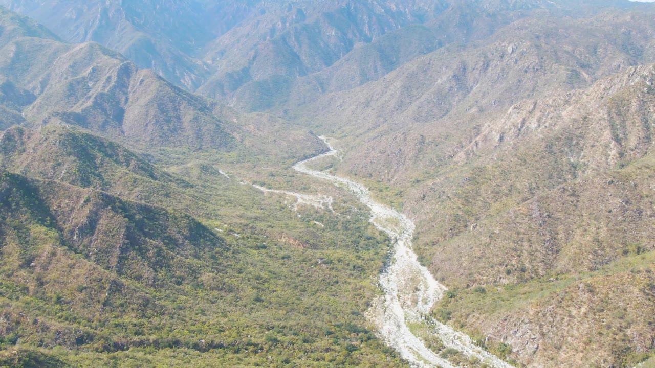 Wide aerial frame from San Dionisio, Baja California Sur, Mexico, with the river channel running from the lower center toward distant blue mountain ridges and no visible people.