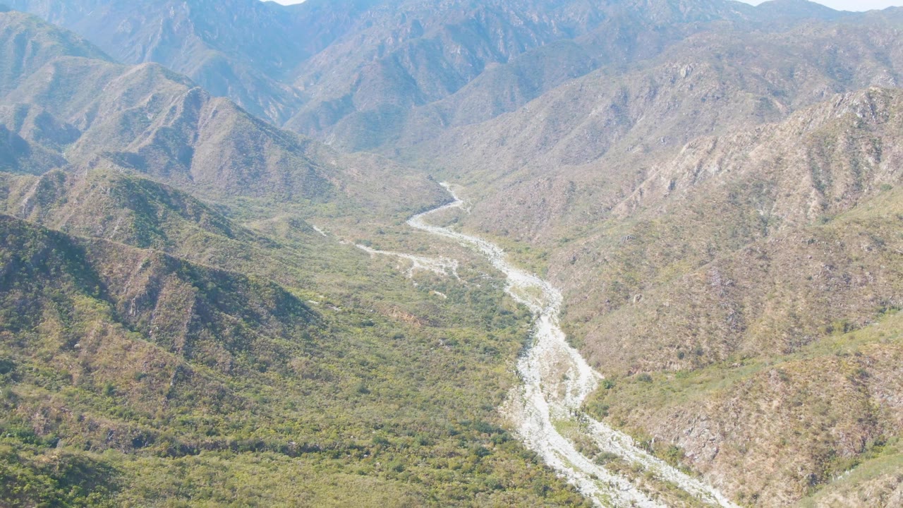 Aerial view over San Dionisio, Baja California Sur, Mexico, where a pale riverbed branches through the center of a green valley below rugged brown hills with no visible people.