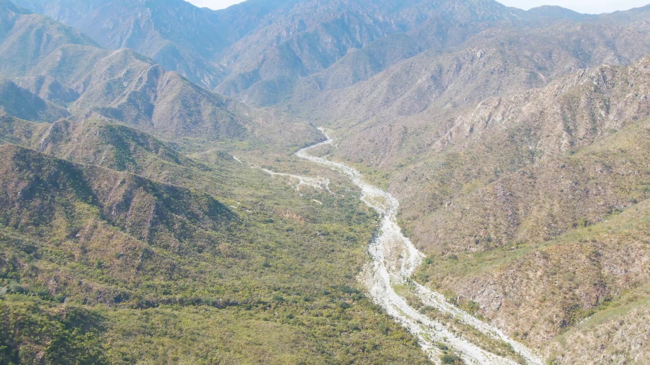 High aerial frame in San Dionisio, Baja California Sur, Mexico, with the bright riverbed curving through the lower valley and steep rocky slopes filling the background, with no visible people.