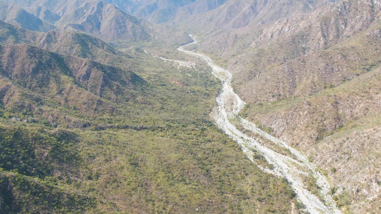 Aerial view in San Dionisio, Baja California Sur, showing a pale winding riverbed crossing green scrub and brown rocky slopes with no visible people.
