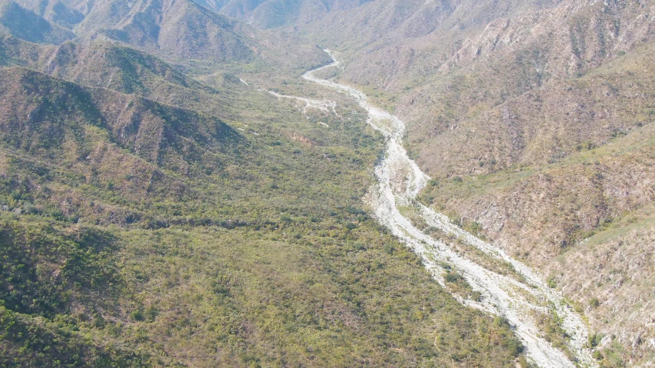 Aerial frame over San Dionisio with the pale riverbed centered between rugged hillsides, green valley vegetation, and no visible people.