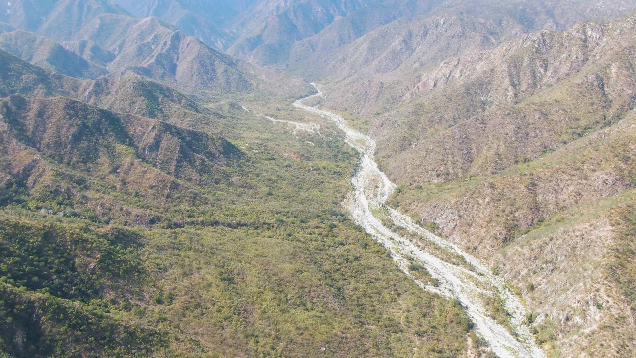 Aerial view of a pale winding riverbed between green valley brush and brown hills.