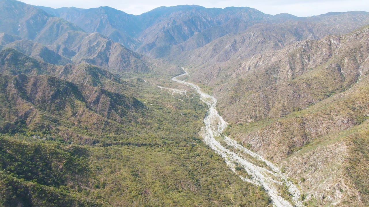 Aerial frame over San Dionisio, Baja California Sur showing a pale riverbed curving through a mountain valley with green-brown slopes and no visible people.