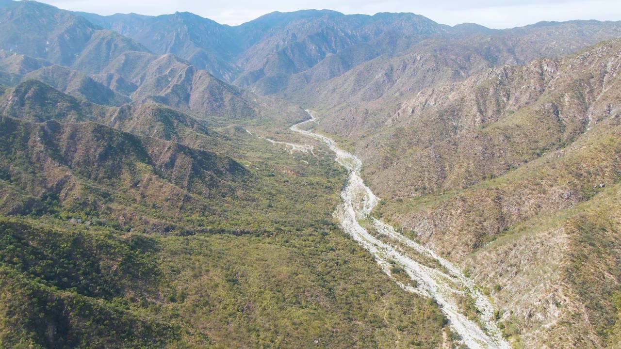 Aerial view in San Dionisio, Baja California Sur with the river channel running from the lower frame toward distant ridges, bordered by rocky hills and no visible people.