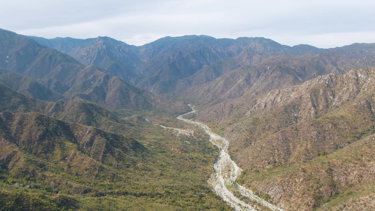 Wide aerial frame over San Dionisio, Baja California Sur, with the river curving through the valley floor between rocky vegetated slopes and no visible people.