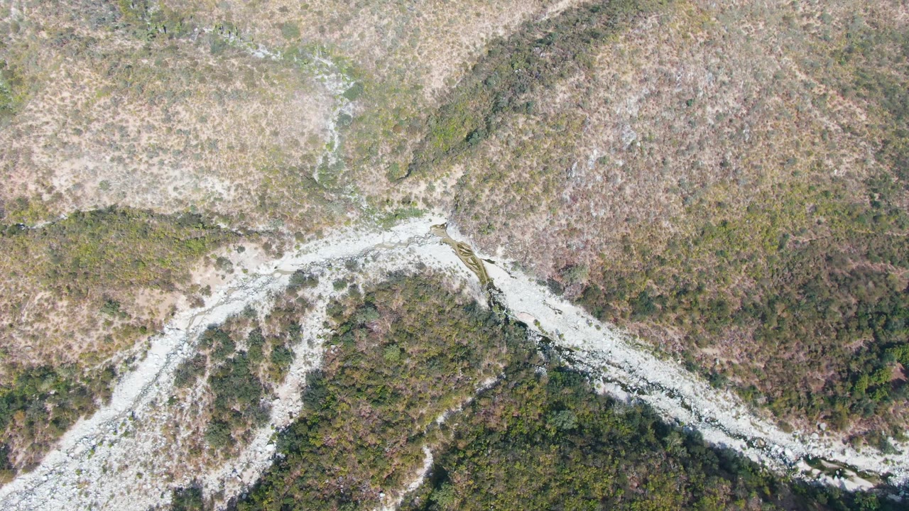 Aerial frame over San Dionisio, Baja California Sur showing a pale winding road or wash crossing brown grasses, scrub, and dark green vegetation, with no visible people.