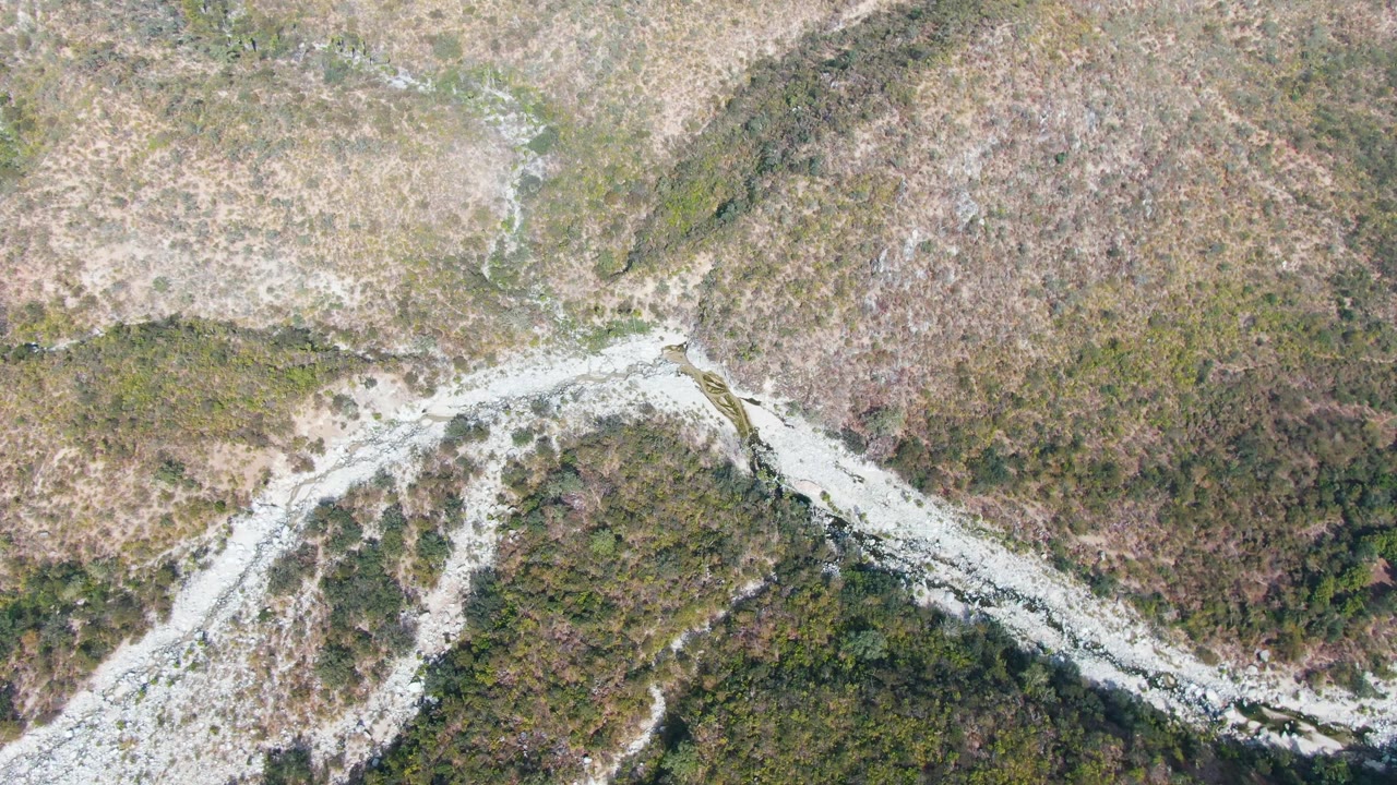 Top-down view in San Dionisio, Baja California Sur with the pale winding road or wash shifting across the center of scrubby brown slope and green forest patches, with no visible people.