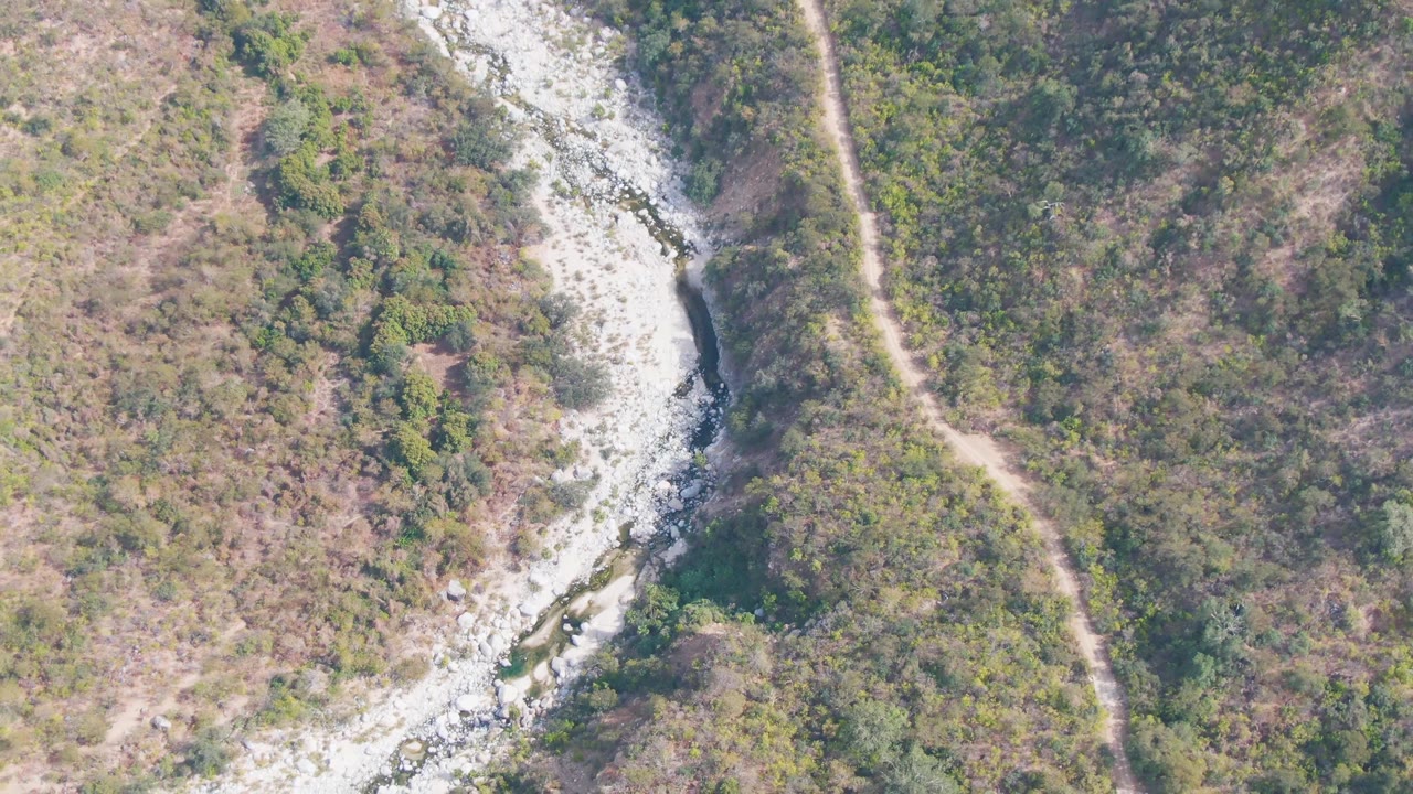 Overhead frame in San Dionisio, Baja California Sur, with the river channel bending down the left side and a thin dirt trail tracing the brushy slope to the right, no visible people.