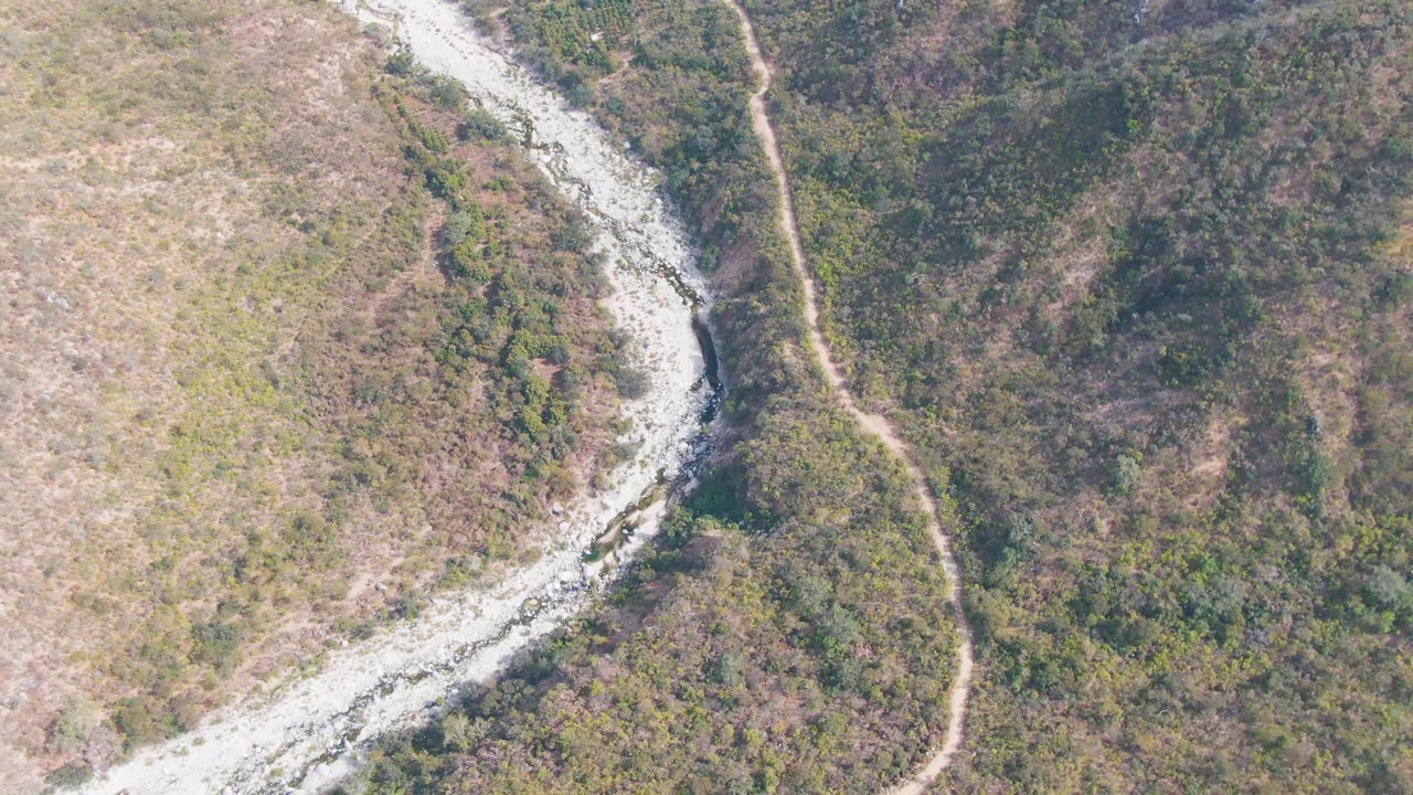 Aerial frame from San Dionisio, Baja California Sur with the pale riverbed bending across the left side, a narrow dirt road near the center-right slope, and no visible people.
