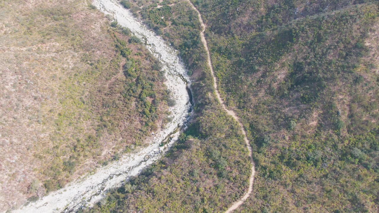 Overhead San Dionisio frame showing the light-toned river channel sweeping down the left of the image, a dirt track along the hill, mixed scrub vegetation, and no visible people.