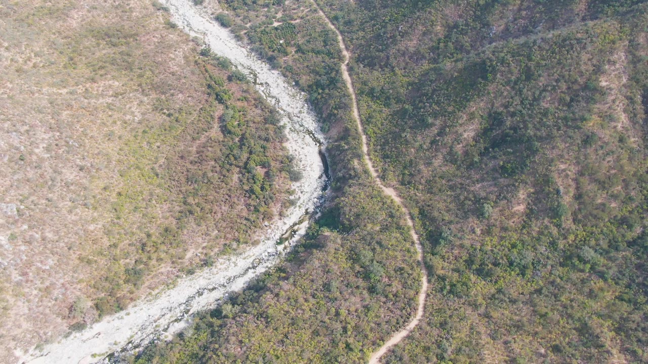 Overhead frame of a winding riverbed crossing dry hills with a dirt road on the slope.