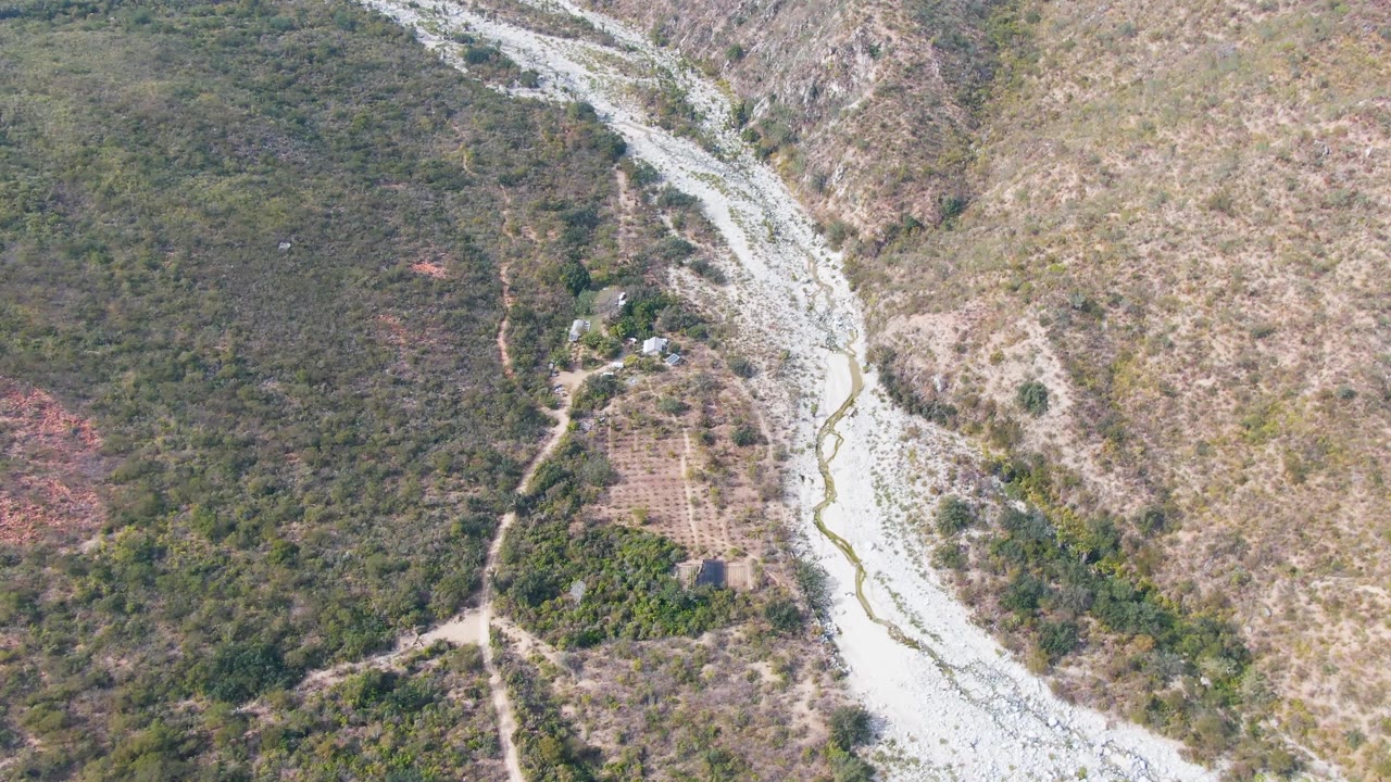 Aerial frame over San Dionisio, Baja California Sur, showing a pale winding riverbed, green scrub slopes, dirt tracks, and small structures near the water with no visible people.