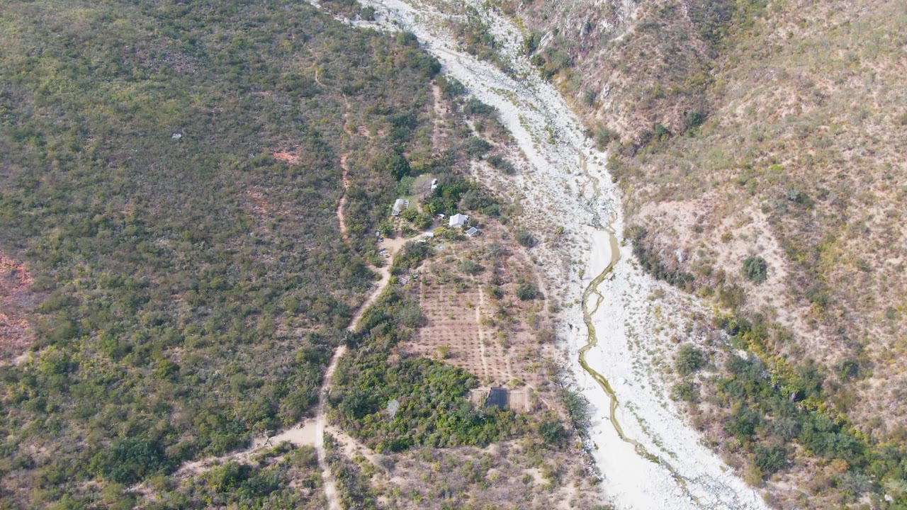 Drone-style frame over San Dionisio showing the riverbed along the right side, scrubby hills, a small group of structures, dirt access tracks, and no visible people.