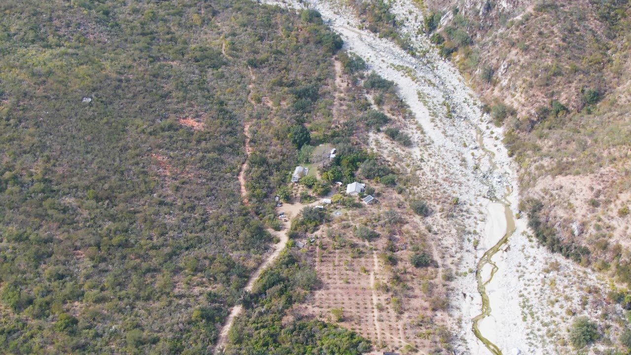 Aerial frame from San Dionisio, Baja California Sur, showing a pale rocky riverbed, thin stream channel, surrounding green-brown hills, and no visible people.