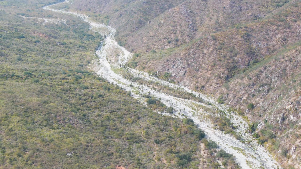 Aerial view over San Dionisio, Baja California Sur, with a pale riverbed curving through green valley vegetation beneath rocky hills and no visible people.