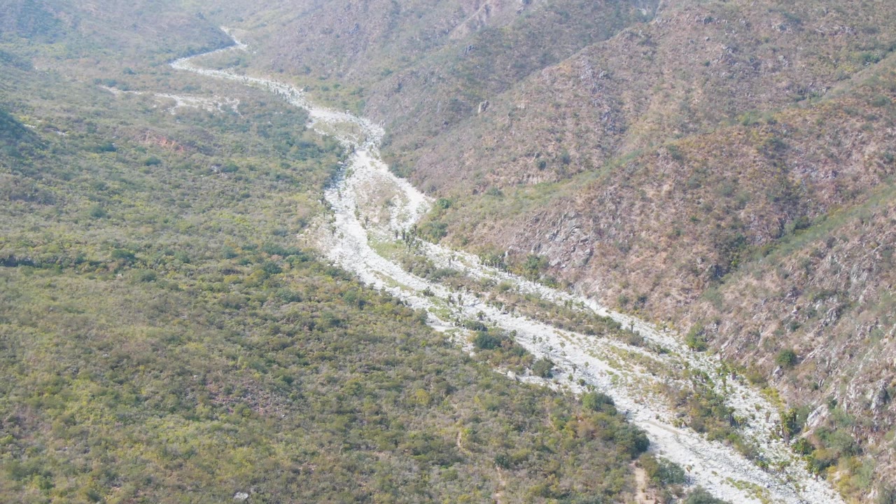 High aerial frame over San Dionisio showing the river channel bending through scrub-covered valley floor, with brown rocky slopes on both sides and no visible people.