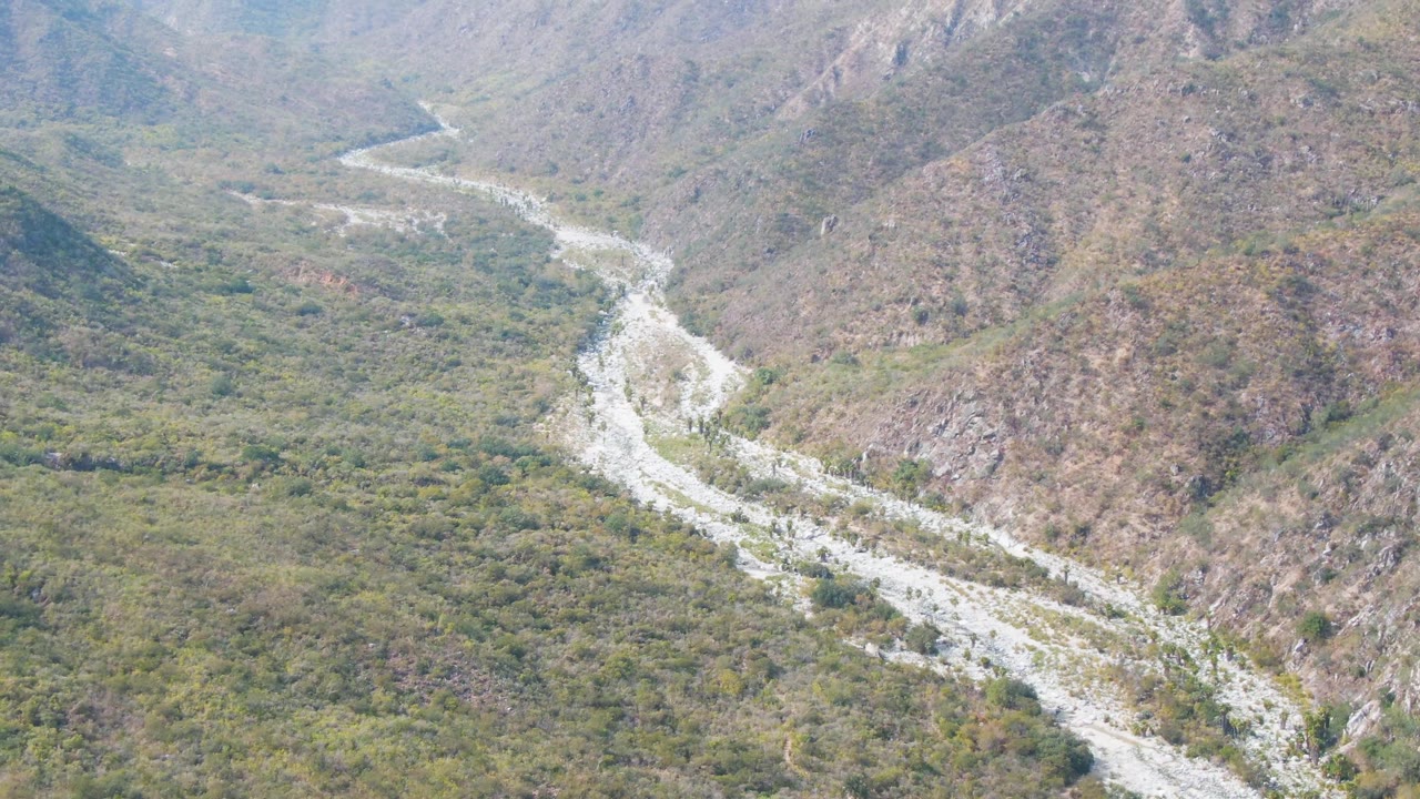 Aerial landscape in San Dionisio with the pale riverbed crossing diagonally through green and brown valley terrain, rocky ridges behind it, and no visible people.