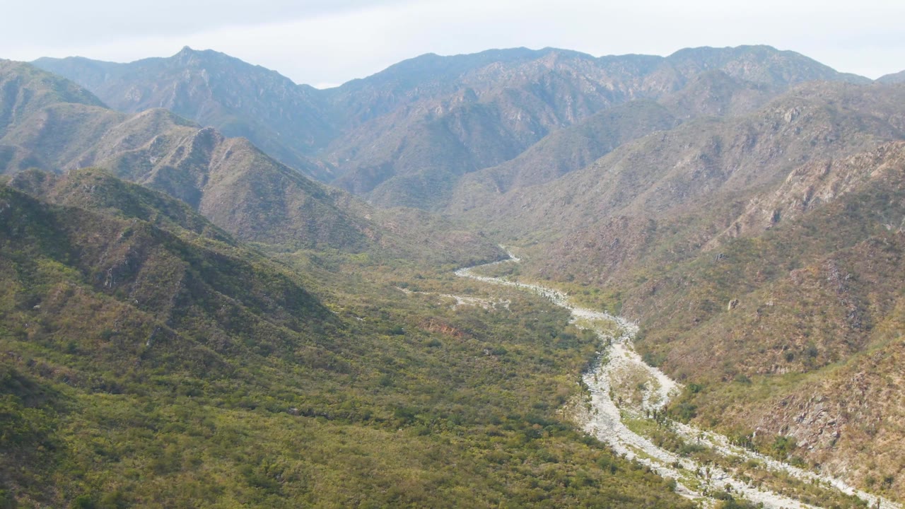 Aerial view over San Dionisio, Baja California Sur, showing a bright river channel crossing the valley floor between brown and green mountains, with no visible people.
