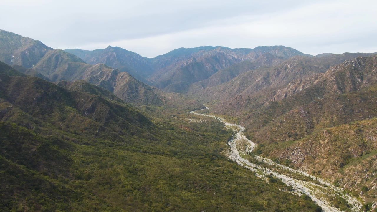 Wide aerial frame of San Dionisio mountain terrain with a pale riverbed curving through green and brown hills, distant ridges, cloud-softened light, and no visible people.