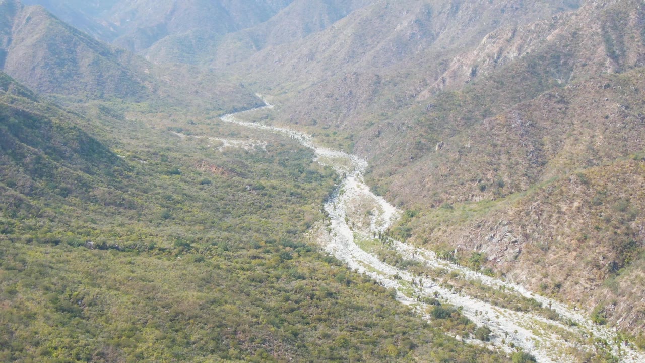 Aerial frame over San Dionisio, Baja California Sur showing a pale winding riverbed entering from the lower left, green valley vegetation, rocky hills, hazy mountain ridges, and no visible people.