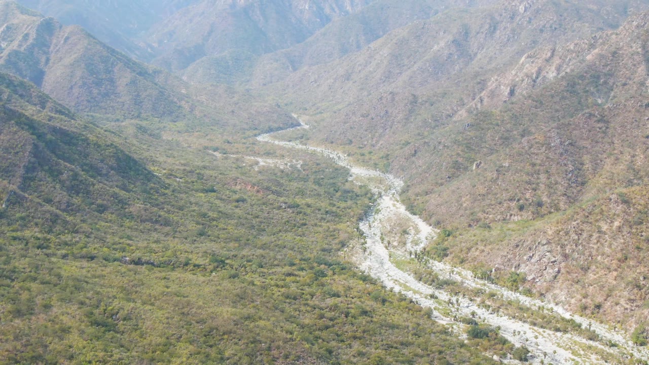 Aerial view in San Dionisio with the pale river wash bending toward the middle distance, steep rocky hills at right and left, low green vegetation across the valley, and no visible people.