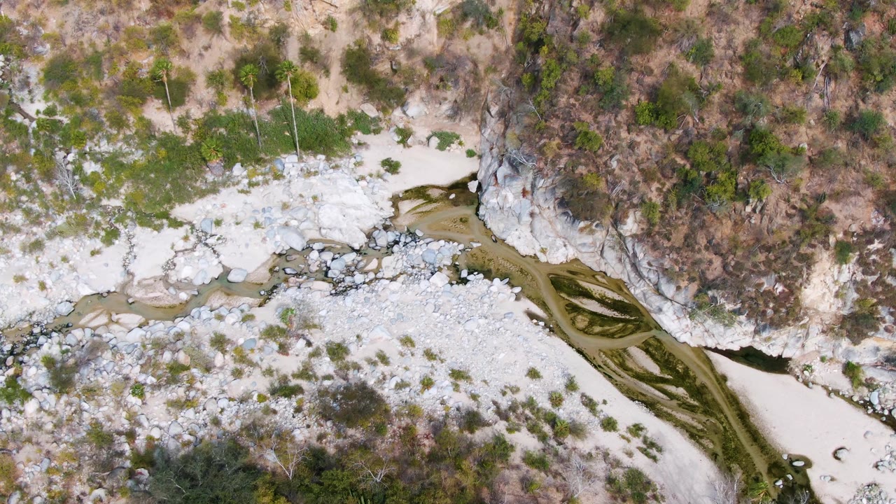 Overhead frame in San Dionisio, Baja California Sur, showing a narrow green-brown river cutting between pale rock beds, dry scrub, and a cliff edge, with no visible people.