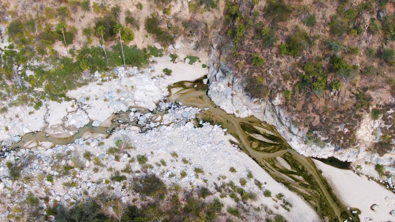 Aerial frame in San Dionisio, Baja California Sur, with the river bending diagonally past white stones, patchy green shrubs, and tan dry ground, with no visible people.