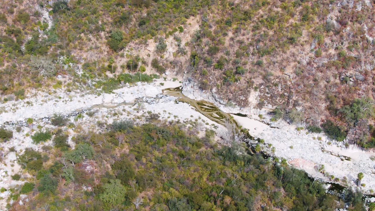 Aerial frame in San Dionisio, Baja California Sur, showing the stream bending through pale stones with brown and green vegetation on the surrounding slopes, no visible people.